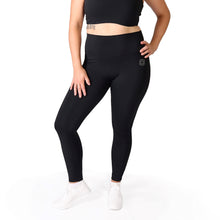Load image into Gallery viewer, Front legs of a woman wearing full length black activewear leggings with a small logo on the left hip below the waistband and white training shoes
