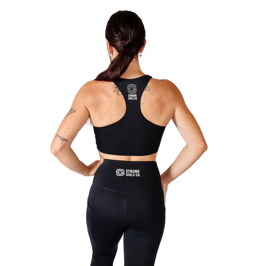 Back facing woman wearing black activewear leggings with a logo on the back middle waistband with the words Strong Girls Co., a matching black crop top with a logo on the back middle upper with the words Strong Girls Co. 