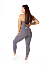 Load image into Gallery viewer, Back facing woman wearing full length grey activewear leggings with pockets with a small logo on the left side ankle, a logo on the back middle waistband with the words Strong Girls Co., a matching grey crop top and white training shoes
